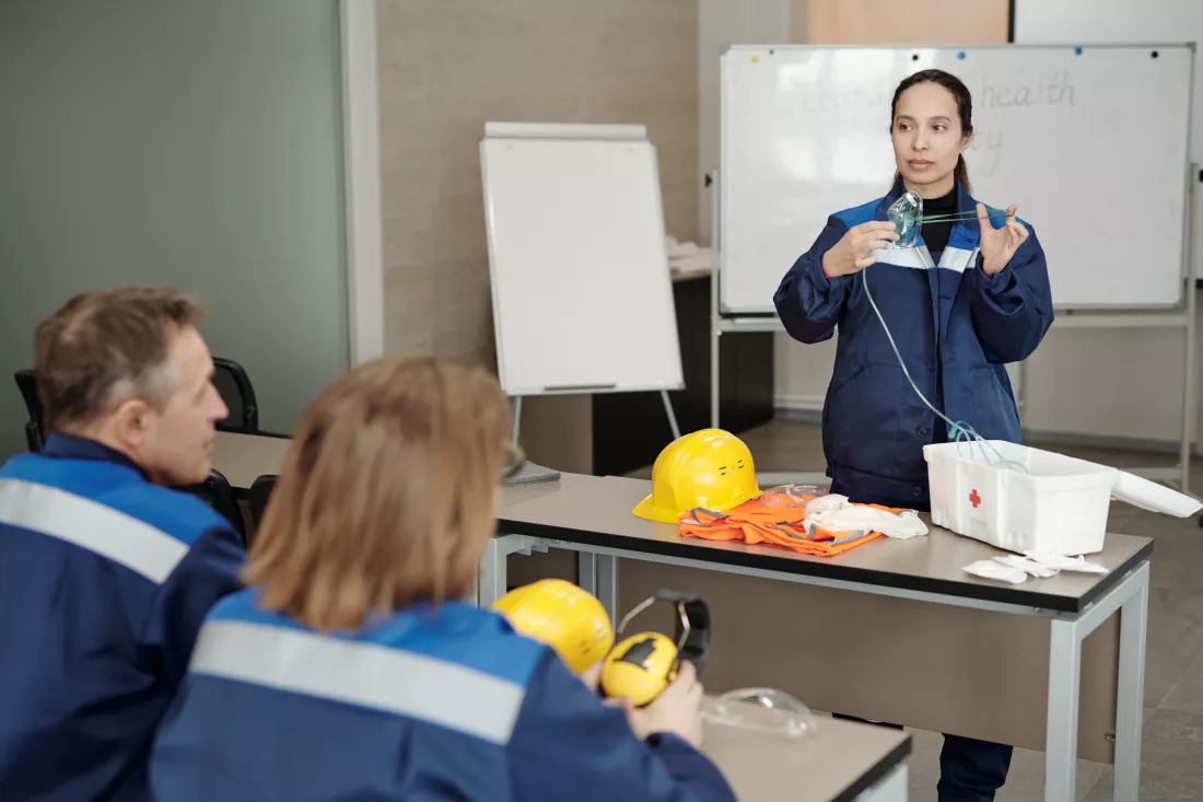 young-female-worker-demonstrating-usage-respirator-while-standing-by-desk-front-colleagues-basics-life-safety-lesson