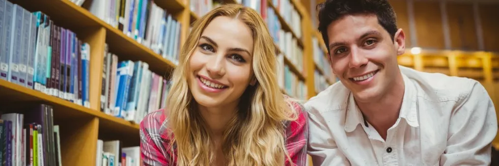 English-Studies-test-smiling-classmates-reading-book-while-leaning-on-bookshelves