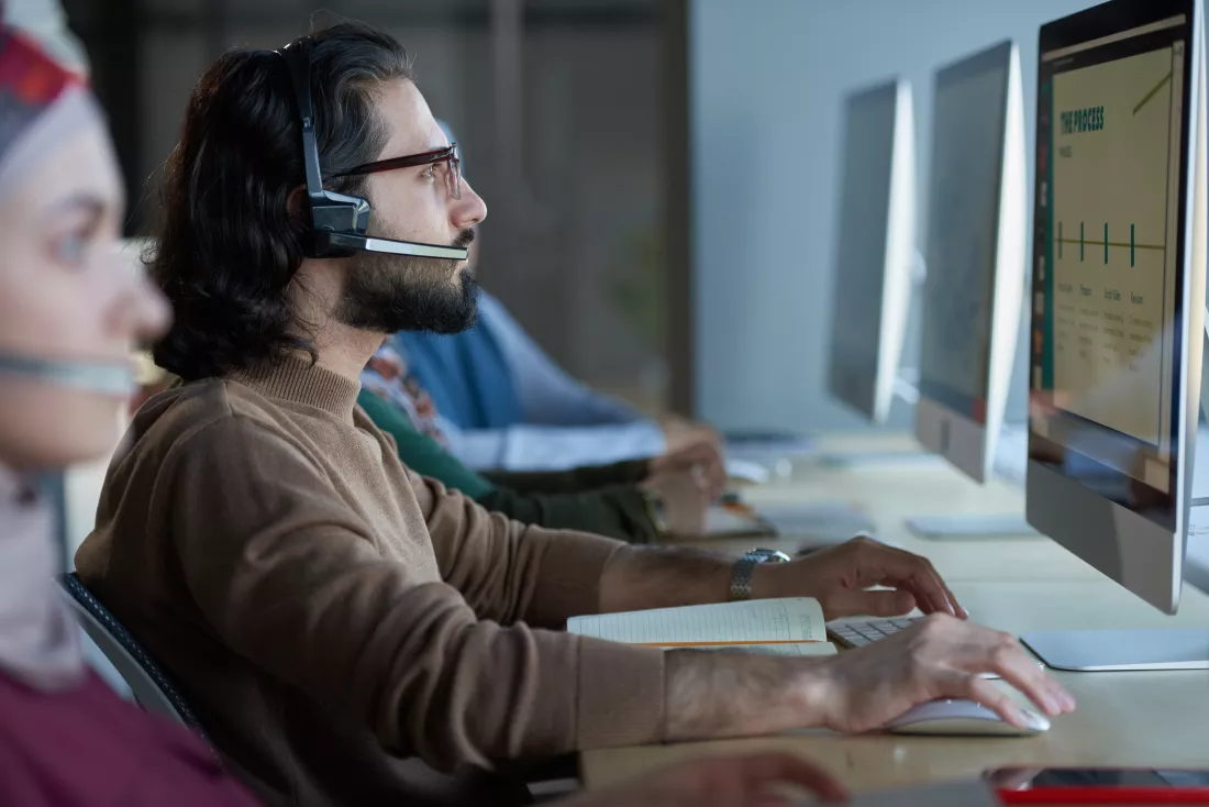 side-view-young-man-headphones-sitting-table-looking-computer-monitor-while-working-call-center